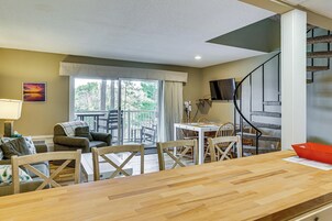 A spacious living area with a wooden dining table, comfortable seating, a staircase, and a large window offering a view of the outdoors.