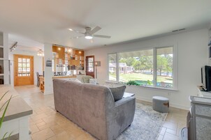 Uma sala de estar espaçosa com um sofá cinzento, área de jantar, cozinha com armários de madeira e uma janela grande com vista para o exterior.