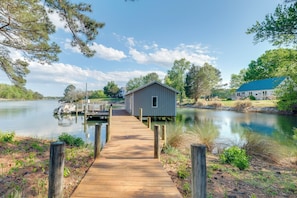A wooden dock leading to a small shed by a calm lake, surrounded by trees and a house with a blue roof. - Fire Pit & Dock! Waterfront Retreat in Heathsville (Heathsville)
