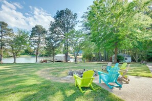 Ein grasbewachsenes Areal im Freien mit Adirondack-Stühlen, einer kleinen Feuerstelle und einem See im Hintergrund.