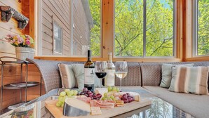 Une pièce chaleureuse au décor intérieur en bois, avec une table en verre sur laquelle sont disposés un plateau de fromages et des bouteilles de vin, et une vue sur les arbres à travers de grandes fenêtres.