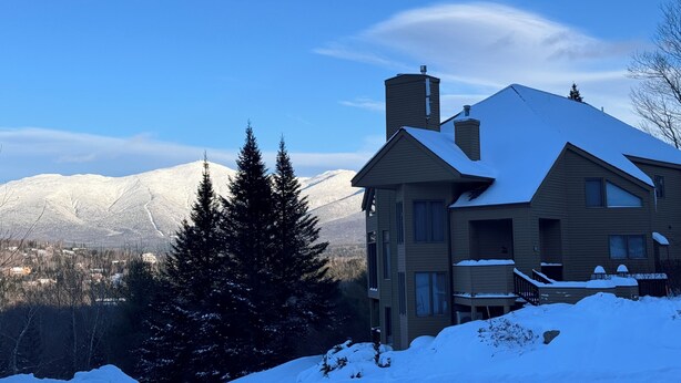 A two-story house with a snow-covered roof and chimney, surrounded by snow and trees, with mountains in the background. - Cr11 C11 Top Rated Townhome Great Views Fireplaces Wifi AC Short Walk to Slopes Pool gym Passes (Bretton Woods)