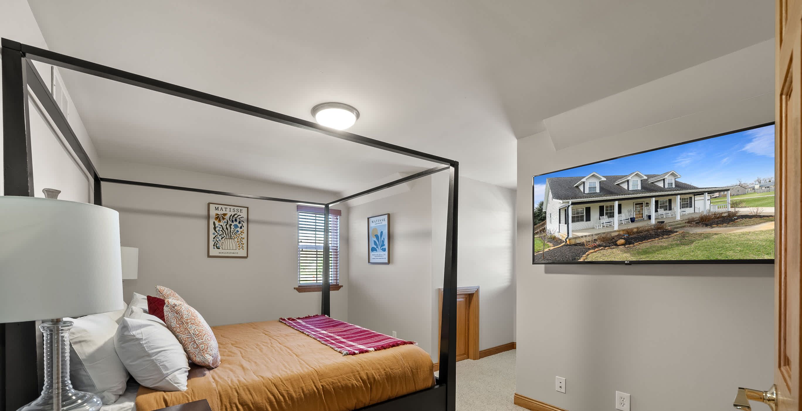 A bedroom with a four-poster bed, a bedside table with a lamp, a framed picture on the wall, and a window with blinds.