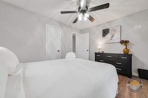 Ein Schlafzimmer mit einem Bett, einem Deckenventilator, einer Kommode, einem gerahmten abstrakten Gemälde und einer Blumenvase.