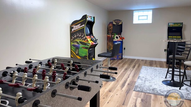 Una habitación con una mesa de futbolín, dos juegos de arcade, un taburete de bar y una ventana.