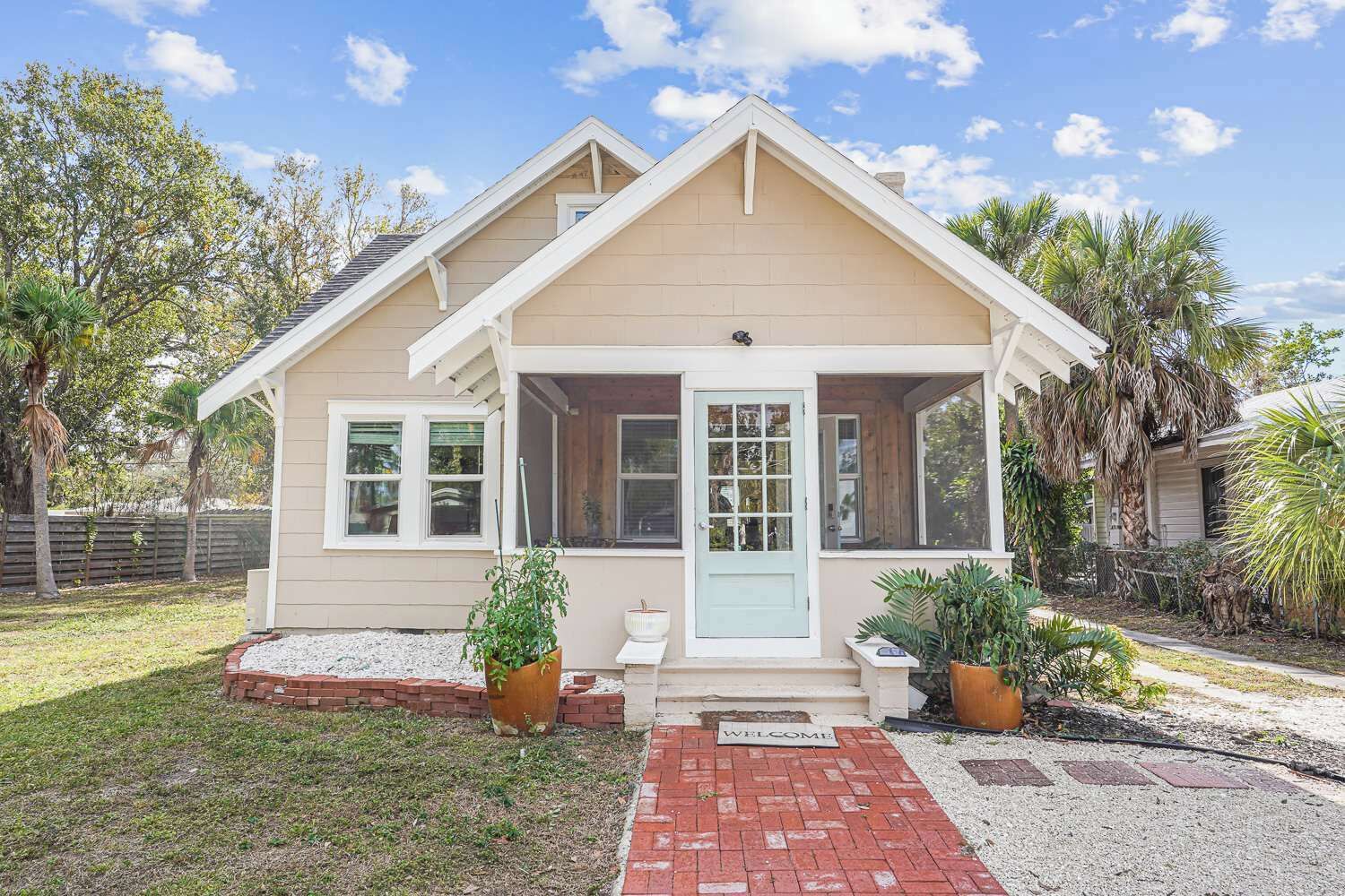A single-story house with a front porch, a small garden area with potted plants, and a welcome mat.