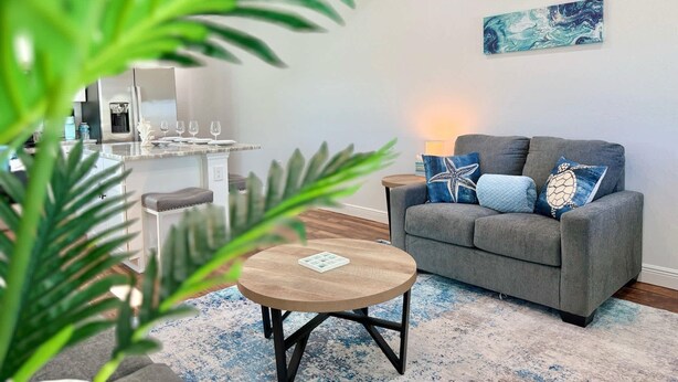 Un salon moderne avec un canapé gris, une table basse ronde en bois et un coin cuisine à l’arrière-plan.
