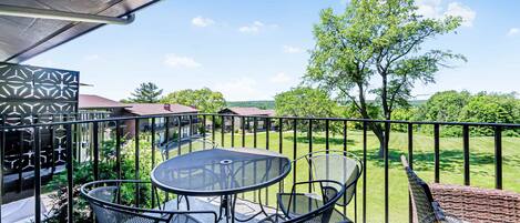 A balcony with a table and chairs, a view of a green lawn, and a building in the background.
