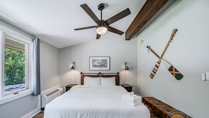 Un dormitorio con cama, ventilador de techo, ventana con cortinas, un cuadro en la pared y dos tablas de paddle surf.
