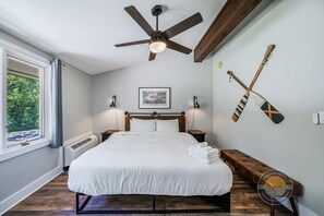 A bedroom with a bed, a ceiling fan, a window with curtains, a wall-mounted picture, and two paddleboards.