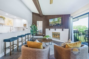 A modern living room with a fireplace, a kitchen with white cabinets, and a balcony with a view.