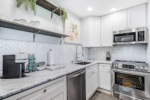 Una cocina moderna con armarios blancos, un microondas de acero inoxidable, horno empotrado, cafetera y salpicadero con motivo de espiga.