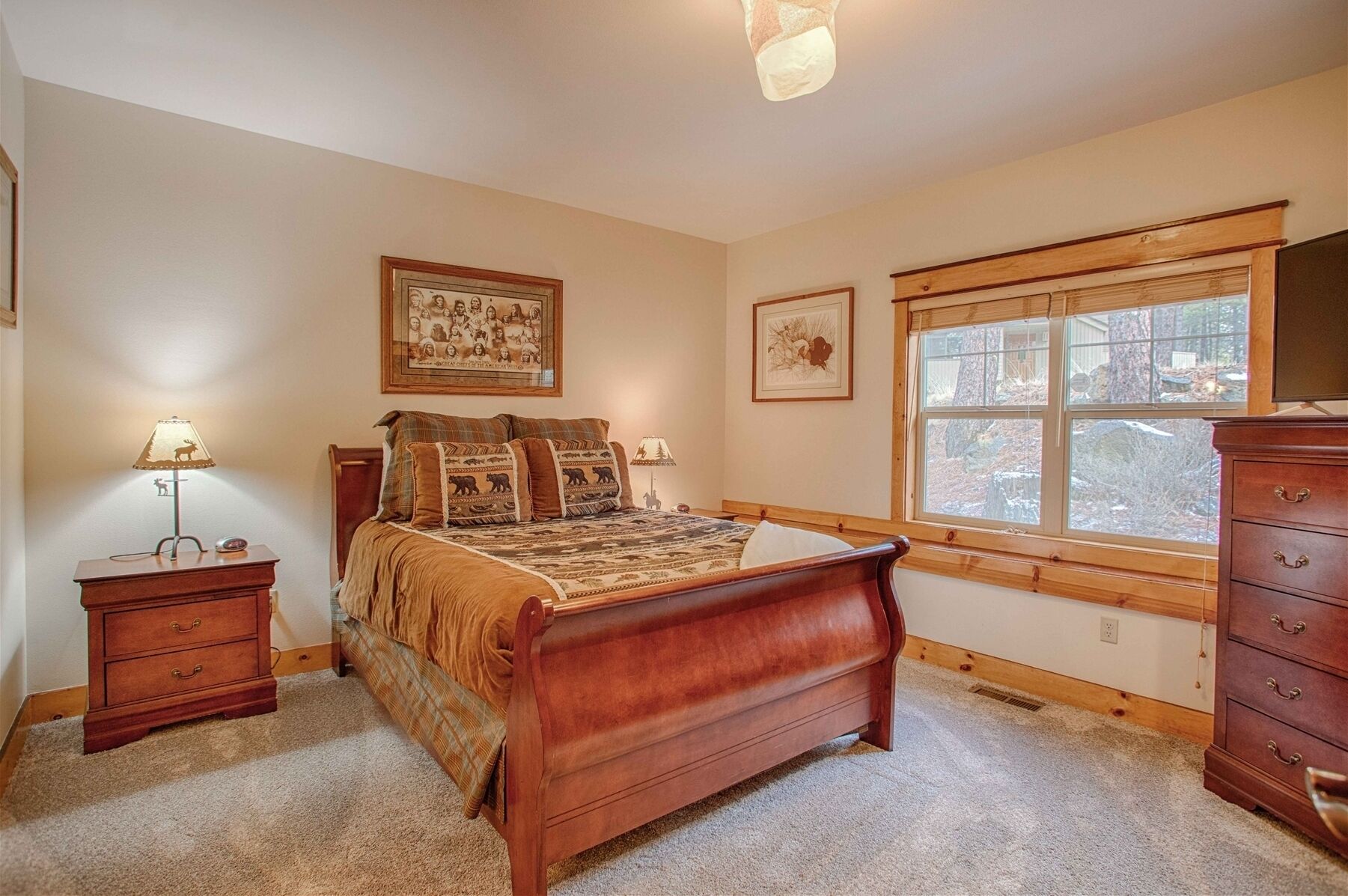 Een slaapkamer met een houten bed, nachtkastje, dressoir en een raam met uitzicht op bomen.