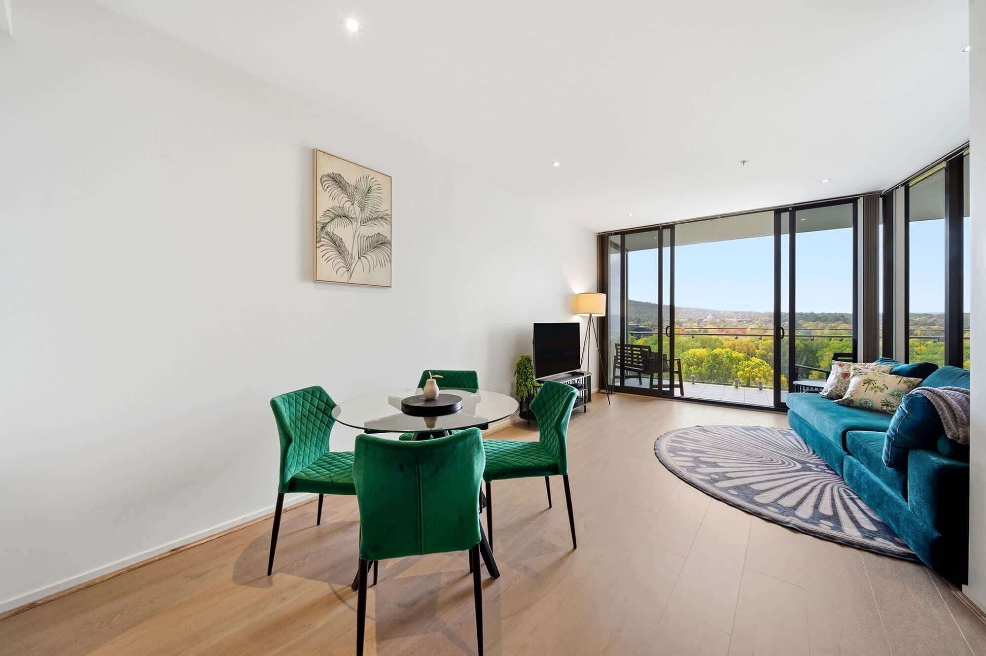 Een moderne woonkamer met een glazen deur naar een balkon, een ronde glazen tafel met stoelen, een bank en een schilderij aan de muur.
