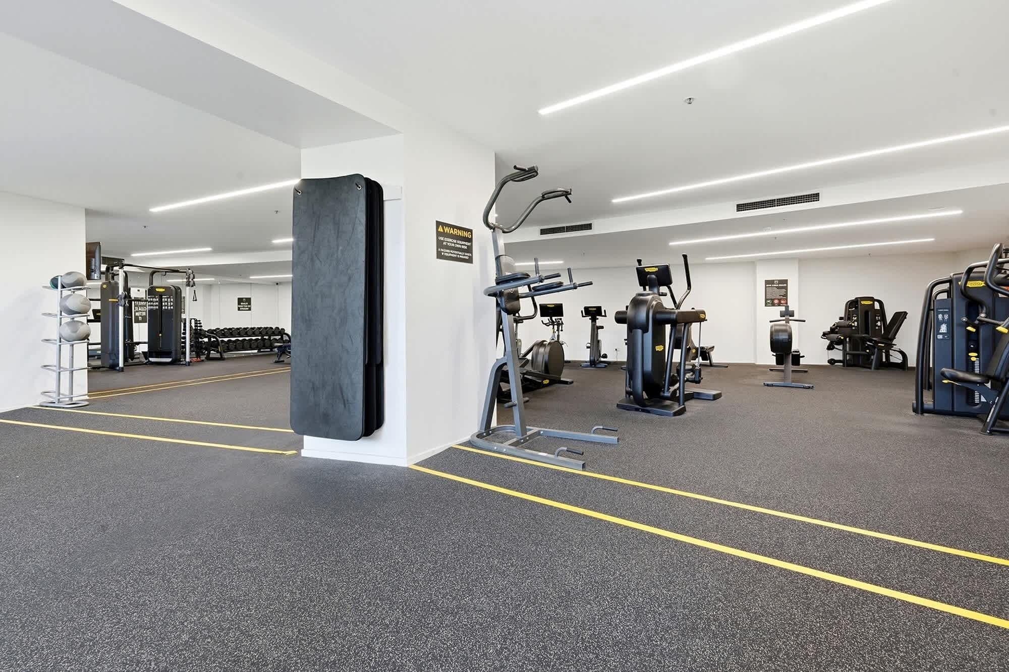 Una palestra con diverse attrezzature per esercizi, un grande specchio e un cartello di avvertimento sul muro.