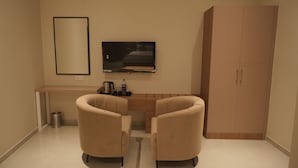 A room with a TV mounted on the wall, a wooden desk, two chairs, a cabinet, and a trash can.