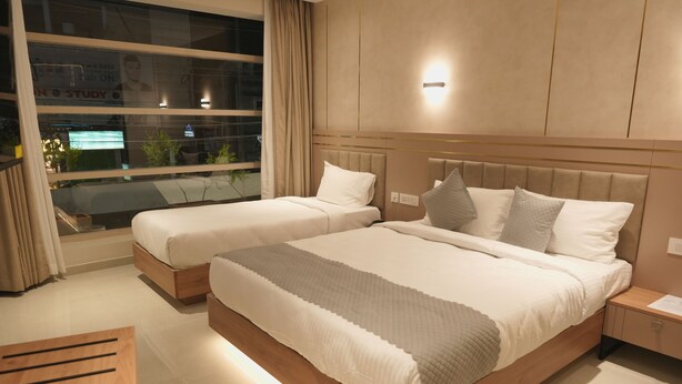 A hotel room with two beds, a nightstand, a wooden side table, and a large window with curtains. - Hotel D' Paris (Kozhikode)