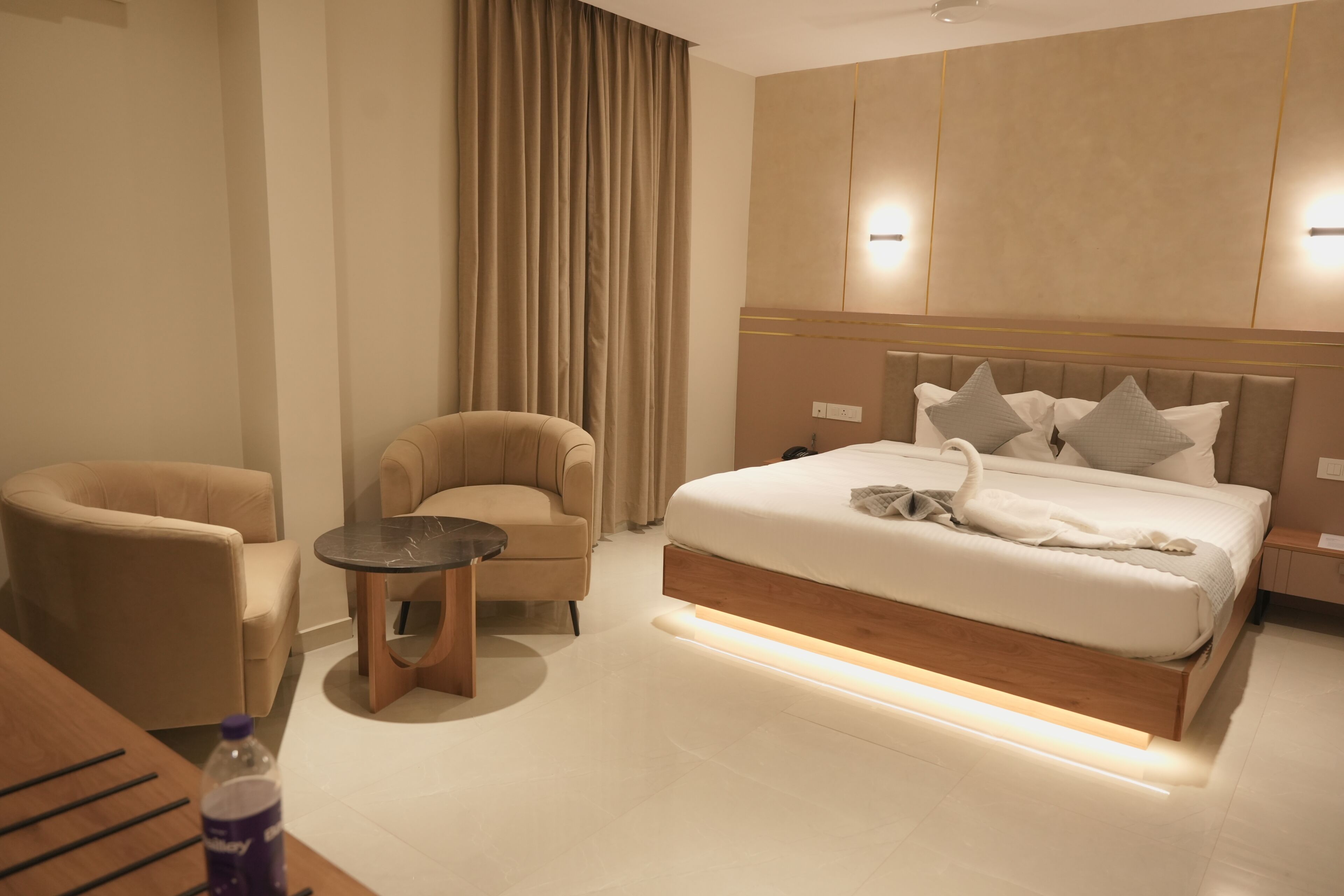 A modern hotel room with a large bed, two armchairs, a small table, and a bottle of water.