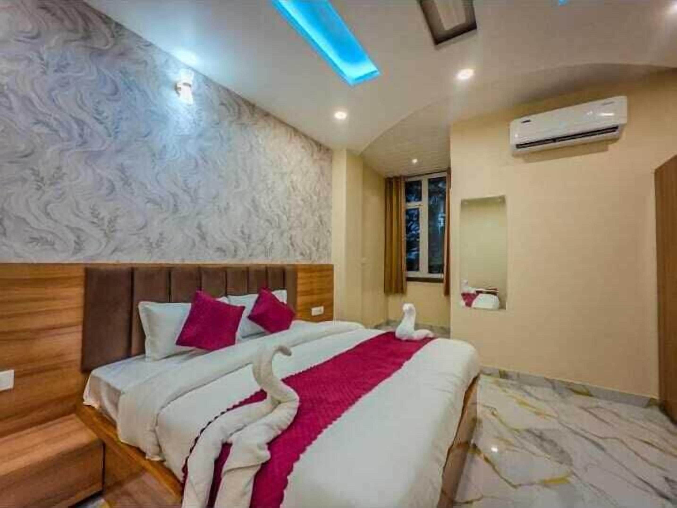 A hotel room with a bed, wooden headboard, patterned wallpaper, an air conditioning unit, and a window with curtains.