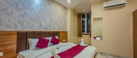 A hotel room with a bed, wooden headboard, patterned wallpaper, an air conditioning unit, and a window with curtains.