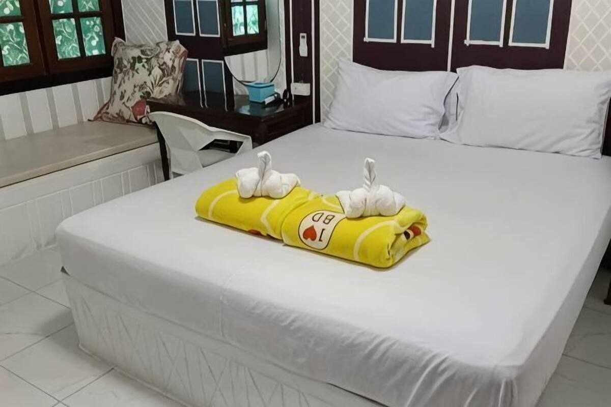 A neatly made bed with a yellow towel and white swan-shaped towels, a white headboard, a window with a view, and a small bedside table with a lamp.