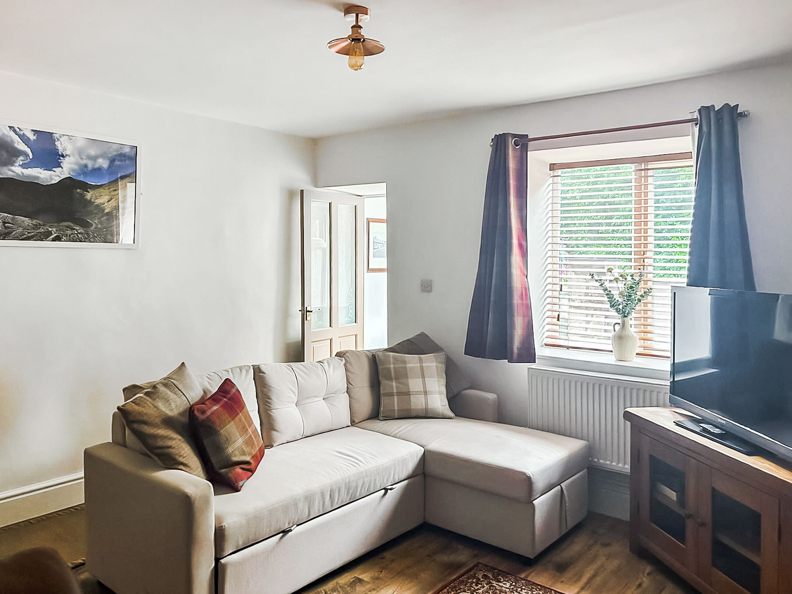 Una sala de estar con un sofá seccional, un televisor sobre un mueble de madera, una ventana con cortinas y una puerta.