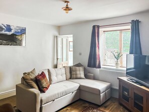 Ein Wohnzimmer mit einem Ecksofa, einem Fernseher auf einer Holzkommode, einem Fenster mit Vorhängen und einer Tür.