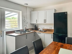 A compact kitchen with white cabinets, a black refrigerator, a wooden dining table, and a window with a view of greenery. - 2 bedroom accommodation in Prestatyn (Prestatyn)
