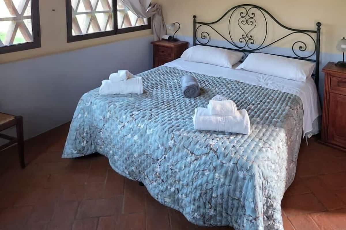 A bedroom with a bed, a nightstand, a window with curtains, a wooden ceiling, and a painting on the wall.