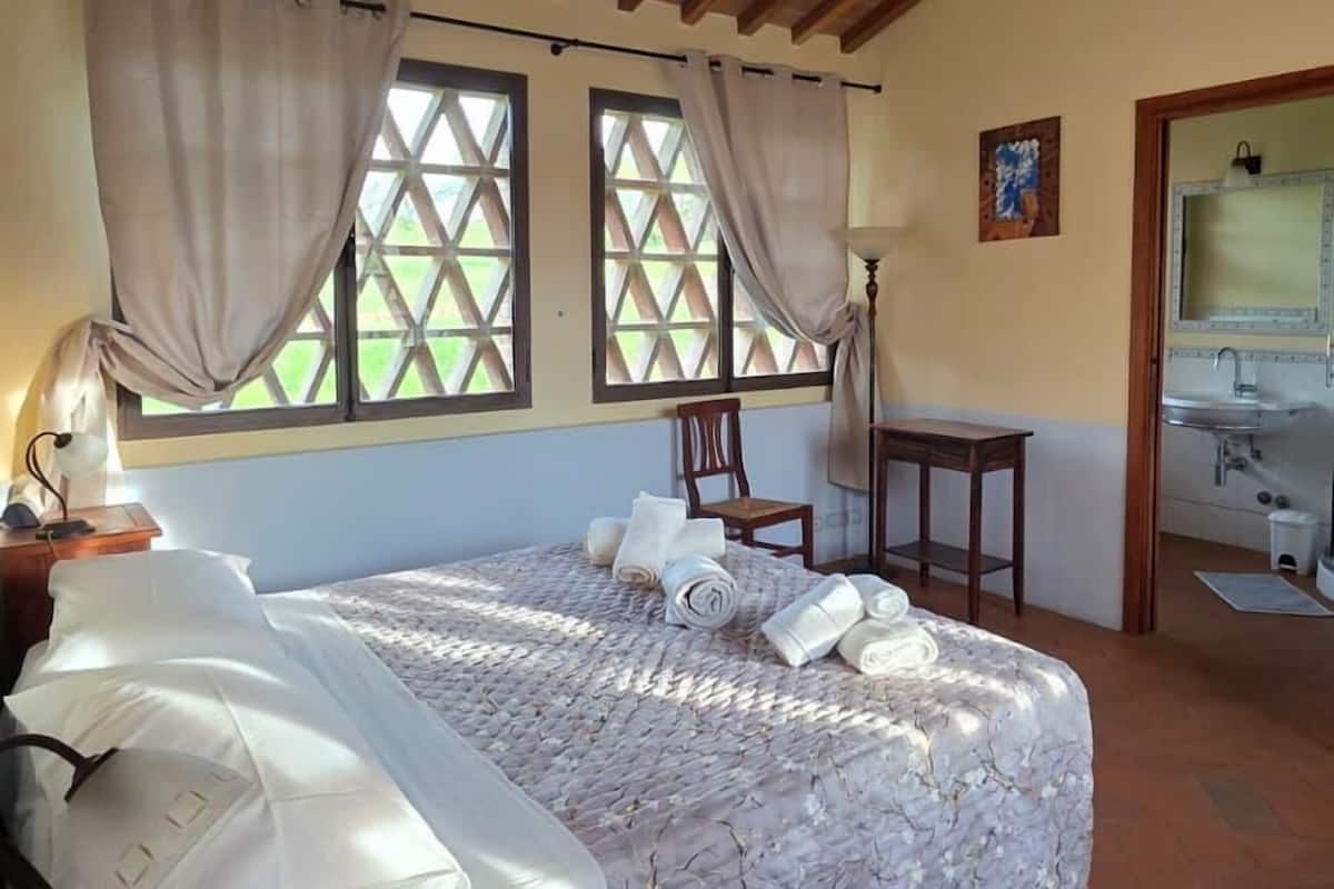 A bedroom with a wooden ceiling, a bed with a patterned bedspread, a chair, a desk, and a window with curtains.