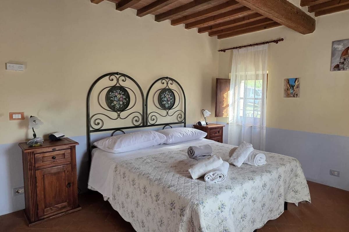A bedroom with a bed, wooden nightstands, a window with sheer curtains, and a wall-mounted painting.