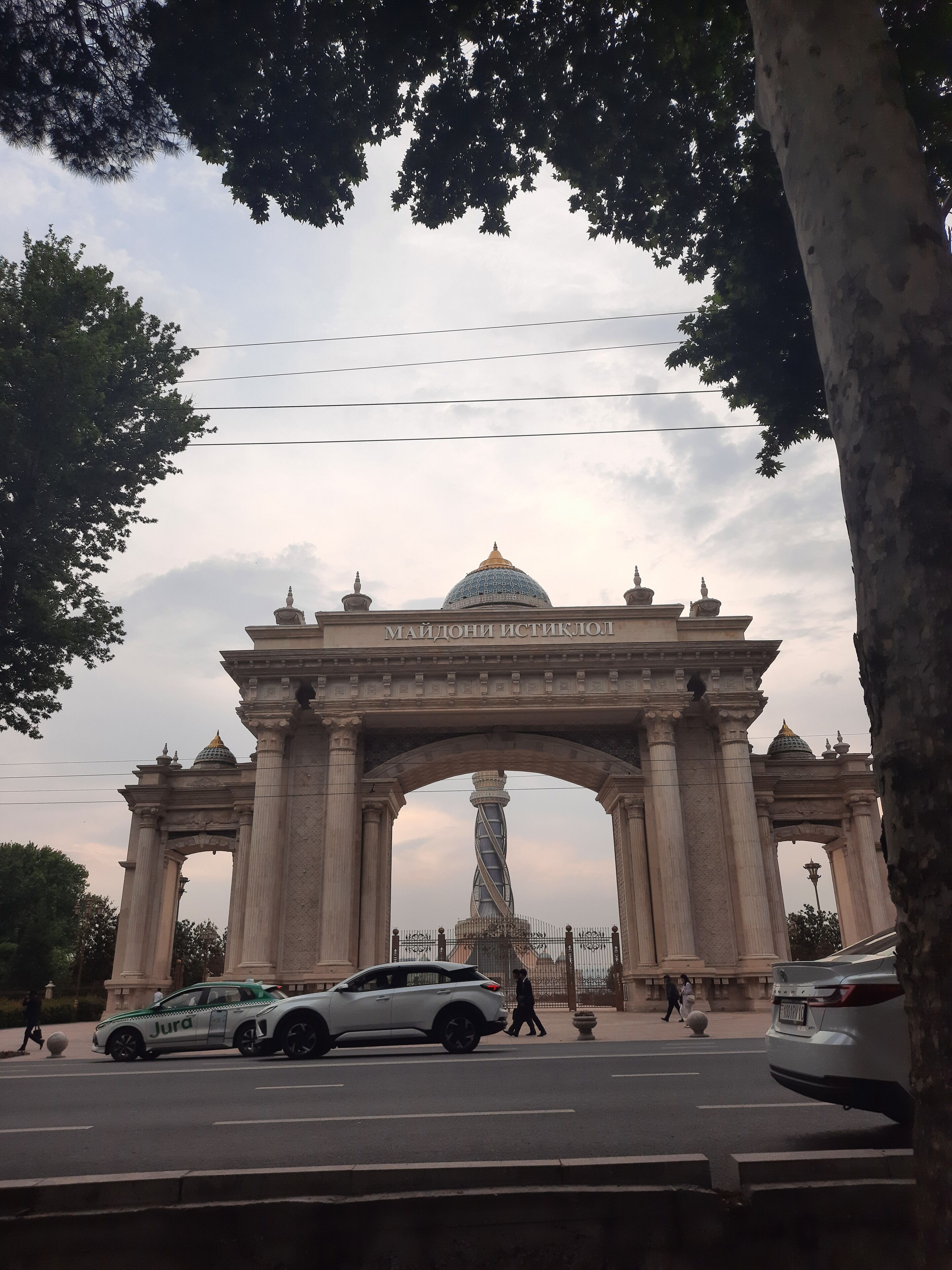 Une arche surmontée d’un dôme et ornée d’inscriptions, entourée d’arbres et bordée par la rue où circulent des véhicules.