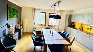 Uma sala de jantar com mesa de madeira, cadeiras azuis, um candelabro e vista para o exterior pelas janelas.