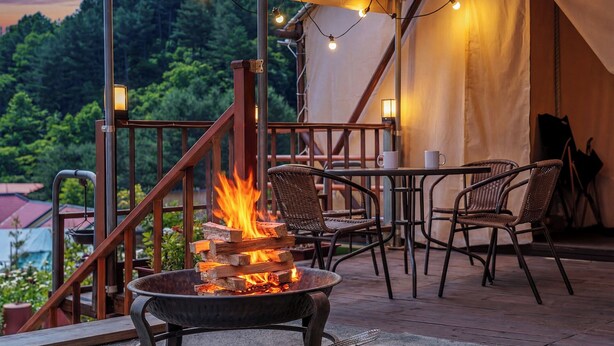 A fire pit with logs, wicker chairs, a table, and a view of a forested area. - Pocheon Terrace 527 Campground (Pocheon)