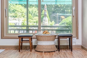 A room with a large window, two wooden stools, a wicker table, and a view of greenery and a statue outside.