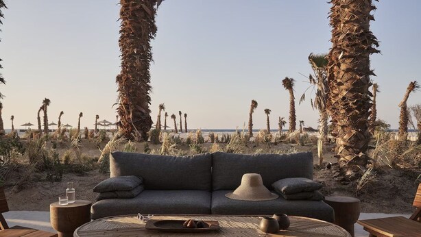 A beachside seating area with a sofa and round table, surrounded by palm trees and a view of the beach. - Casa White (El Alamein)
