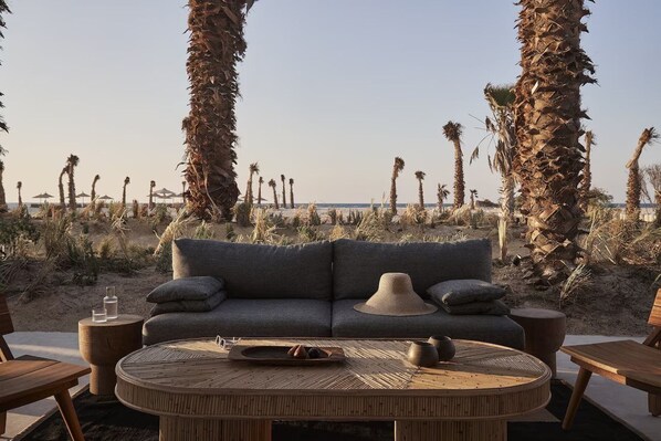 A beachside seating area with a sofa and round table, surrounded by palm trees and a view of the beach. - Casa White (El Alamein)