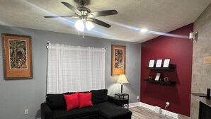 Ein Wohnzimmer mit einem Ecksofa, Deckenventilator, einer Lampe, gerahmten Kunstwerken und einem Kamin.