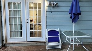 Eine Terrasse mit einem weißen Tisch, einem blauen Stuhl, einem blauen Sonnenschirm und einer weißen Tür mit Fensterfeldern.