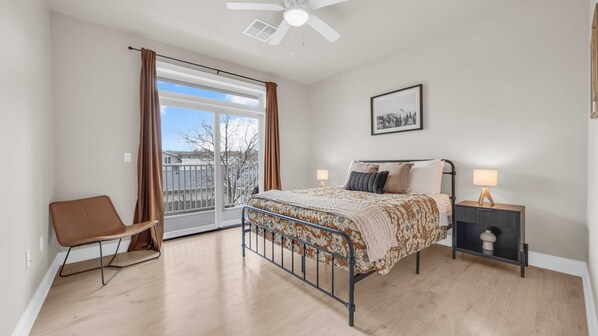 A bedroom with a bed, a chair, a nightstand, a ceiling fan, and a window with curtains.