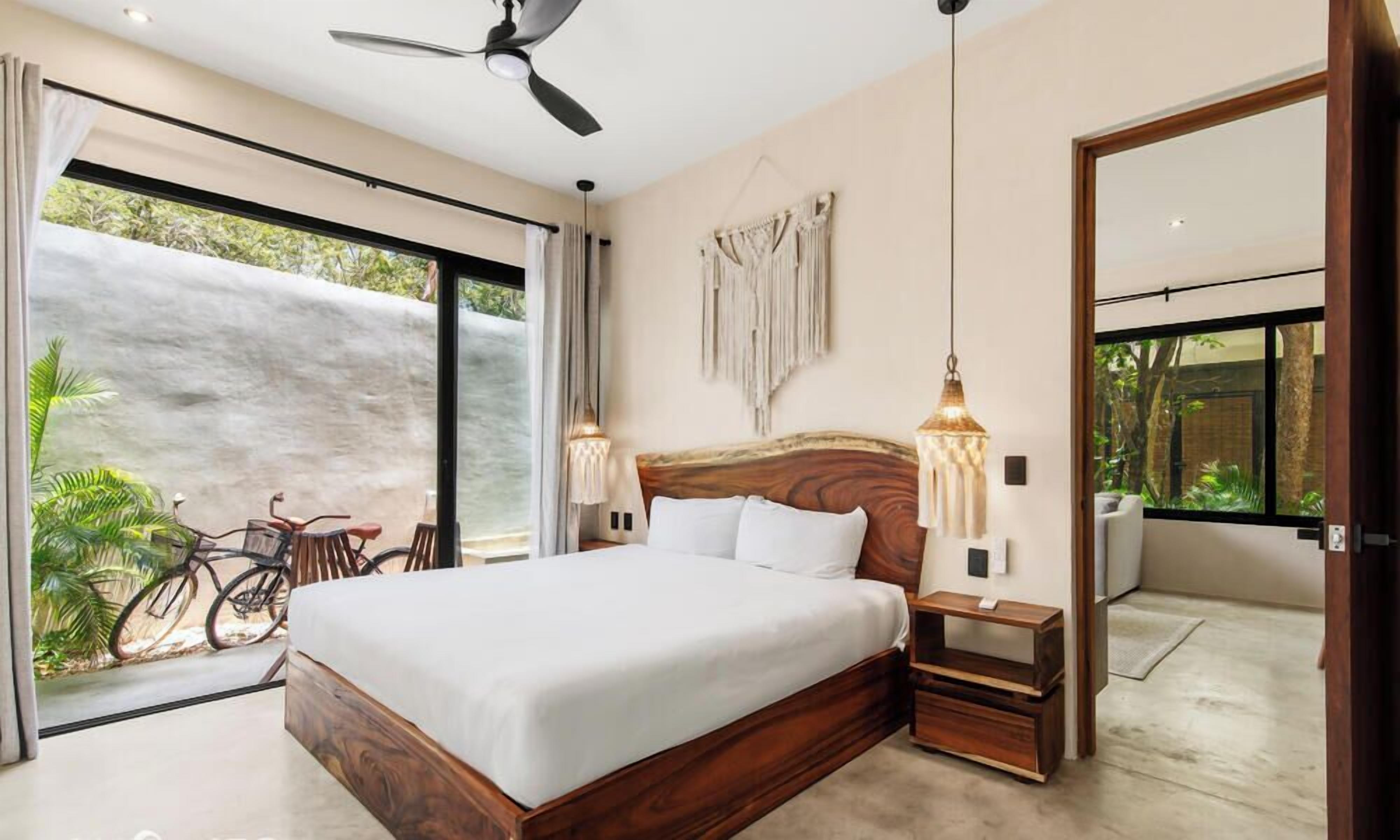 A bedroom with a large bed, a ceiling fan, a nightstand, a wall-mounted lamp, and a view of the outdoors.