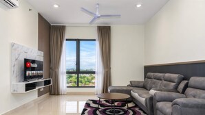 A modern living room with a grey sofa, a TV on a marble shelf, a ceiling fan, and a view through a large window. - Chic 2BR Stay by Jonker Street (Malacca City)