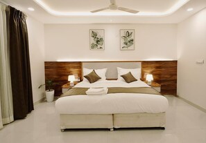 Quarto de hotel moderno com uma cama grande, duas mesas de cabeceira com abajures, um ventilador de teto e duas gravuras botânicas emolduradas na parede.