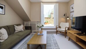 Una sala de estar con un sofá, una mesa de centro, un televisor, una silla y un ventanal con vistas a un jardín.