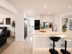 A modern kitchen with white cabinetry, a central island, and a bar area with stools. - Côte d’Azur - 4BR GC Waterfront Dream Home with Pool (Broadbeach Waters)