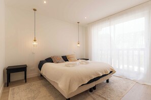 Ein Schlafzimmer mit einem Bett, Nachttischen und einem großen Fenster mit durchscheinenden Vorhängen.