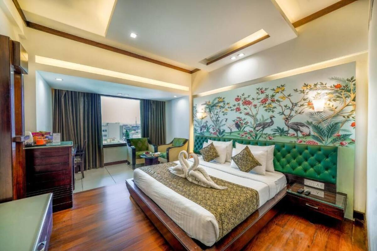 Une chambre d’hôtel avec un grand lit, une tête de lit recouverte d’un tissu vert, une fresque murale aux motifs floraux et ornithologiques, un bureau avec une chaise et une vue par la fenêtre.