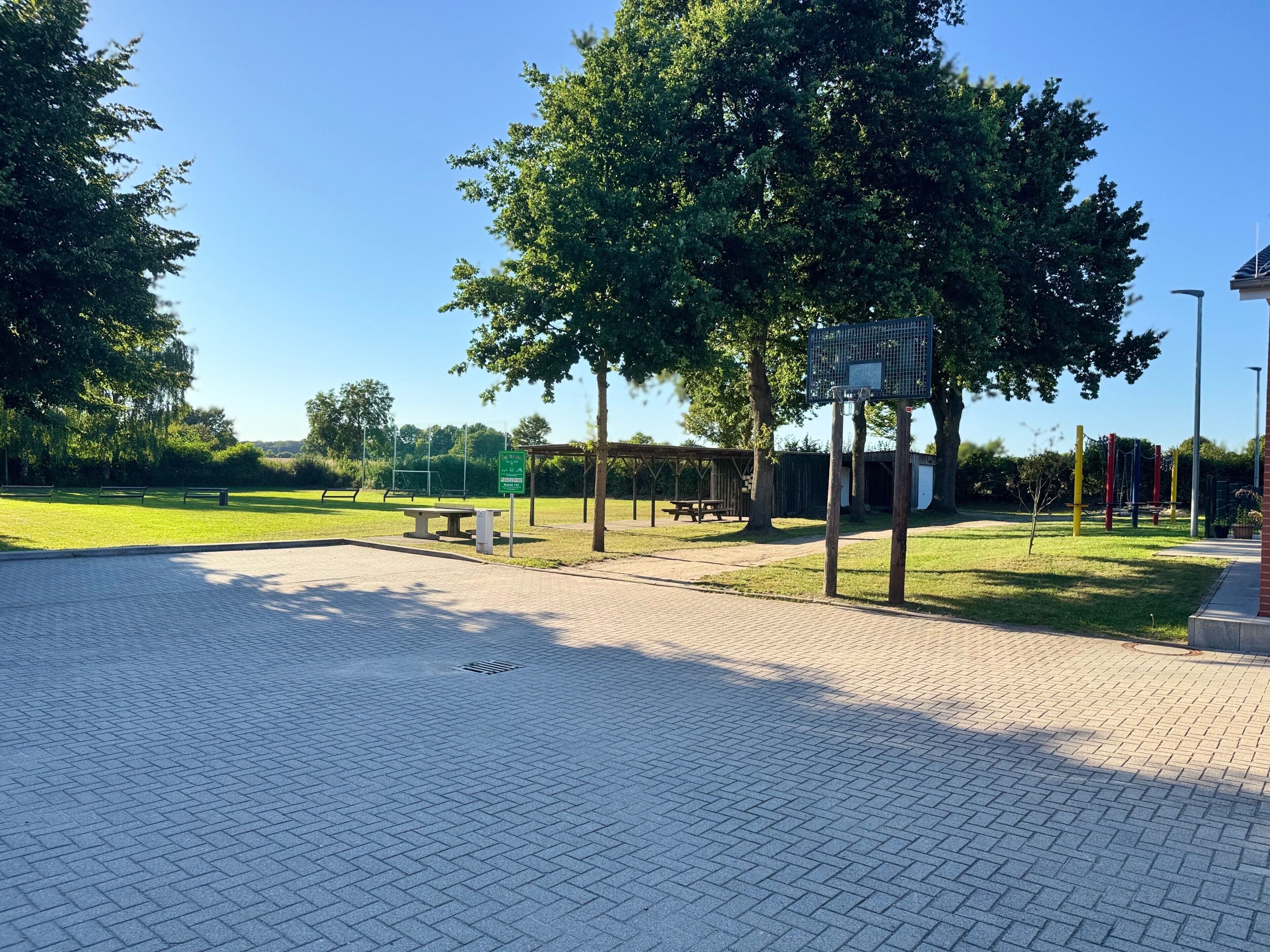 A paved outdoor area with a basketball hoop, picnic tables, and a playground.