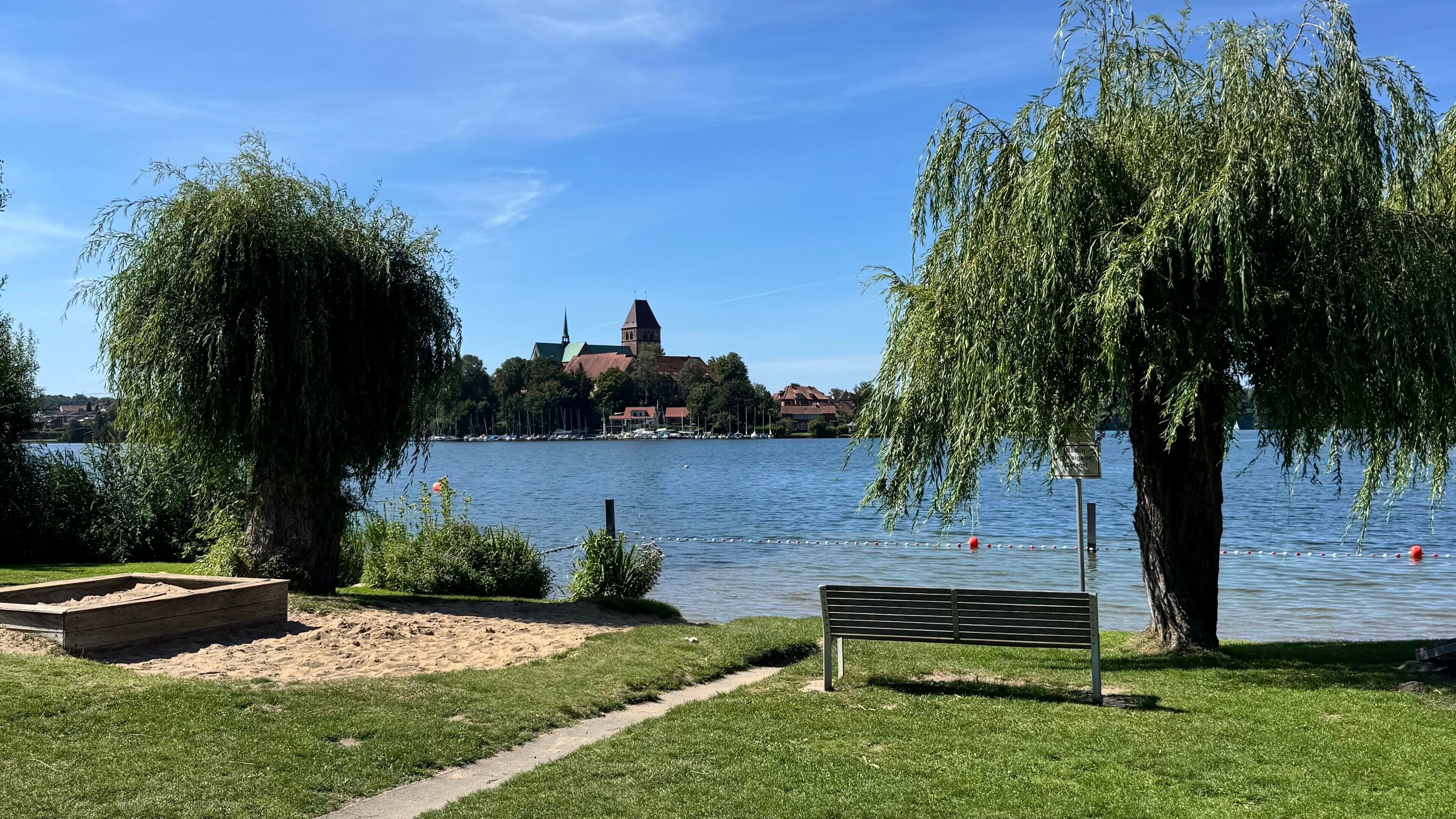 A lakeside park with a bench, trees, and a distant building with a tower.