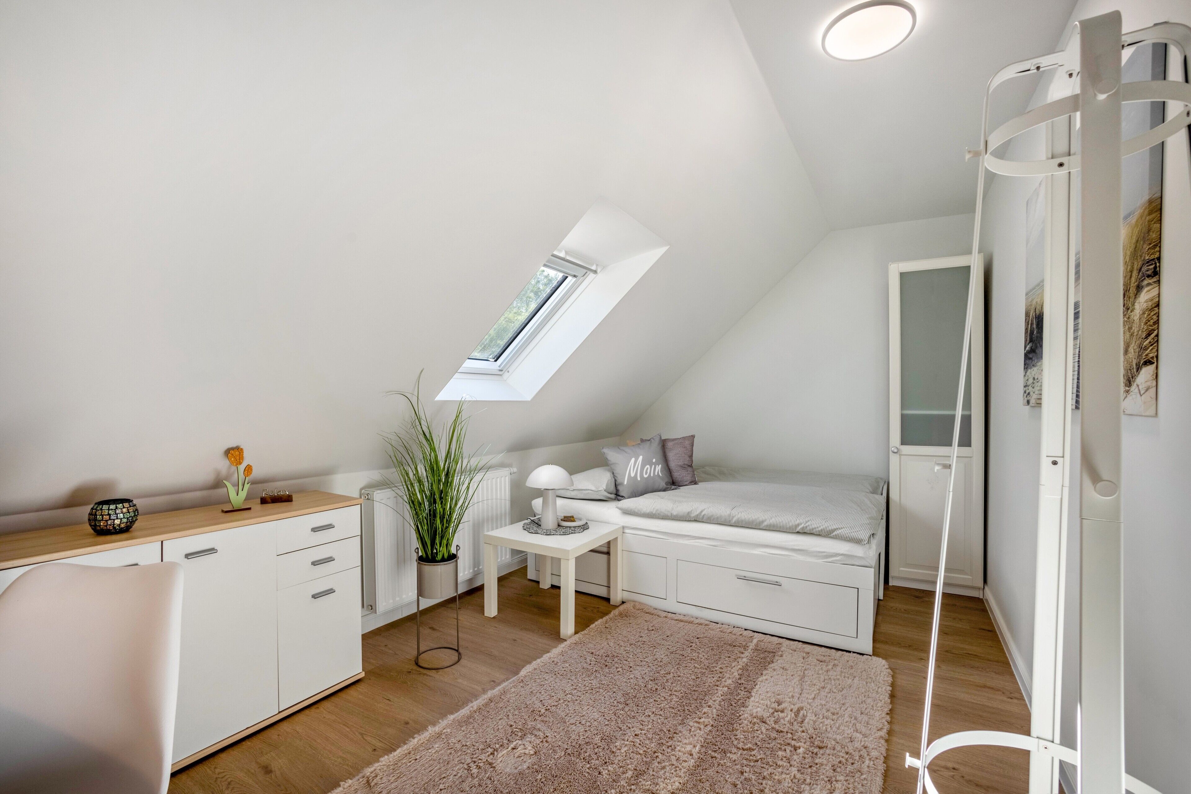 Une pièce compacte et bien éclairée, avec un plafond en pente, une lucarne, un lit, une petite table et un meuble blanc.